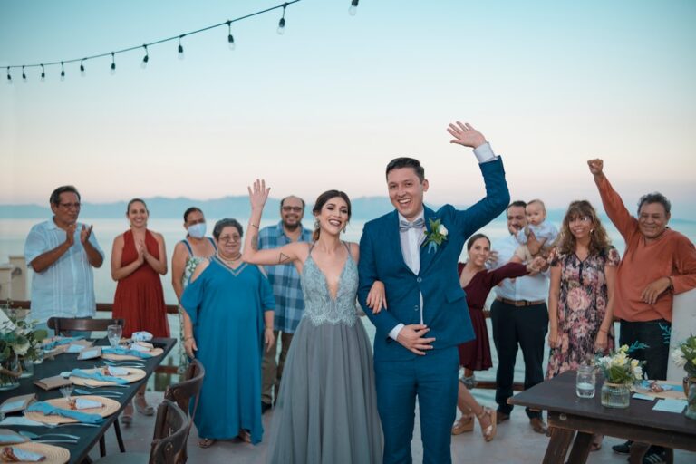A couple shows an engagement ring to smiling family gathered in a cozy living room.