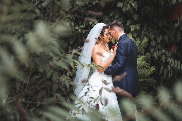 A couple poses for wedding photos outdoors with mountain scenery and friends gathered nearby.